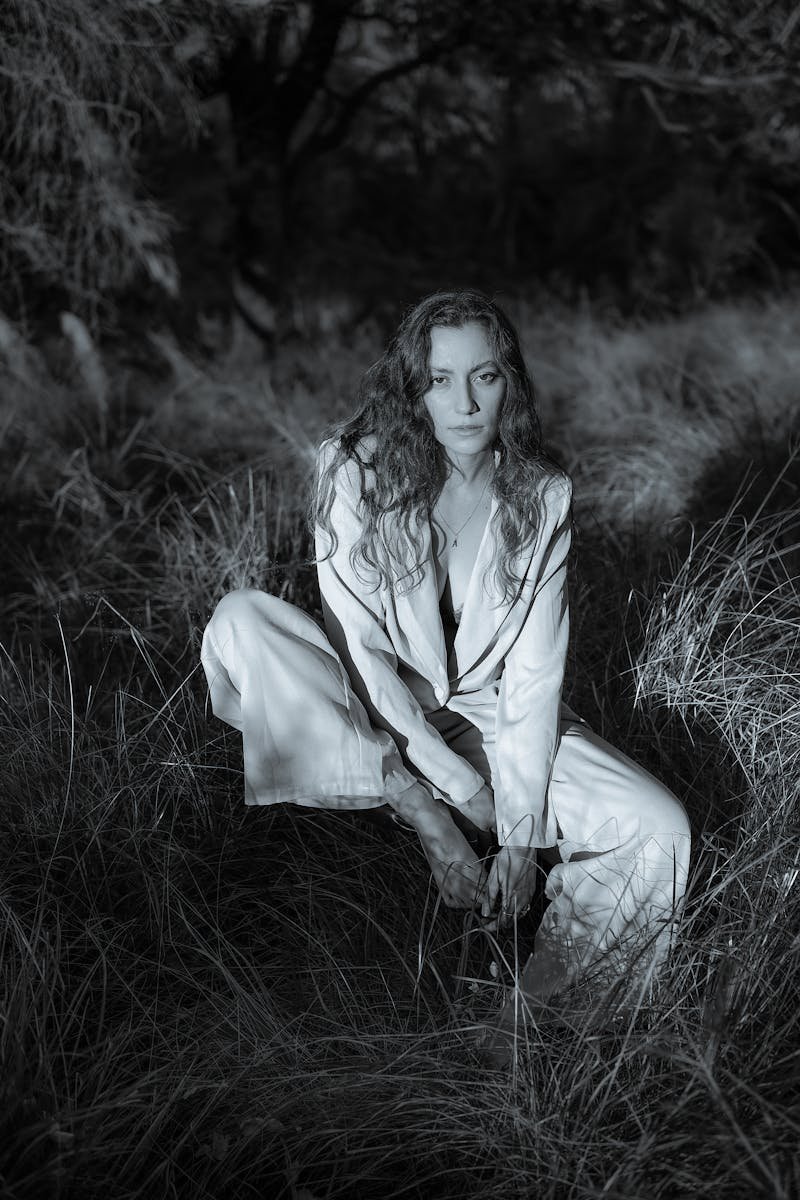 A woman kneels in grass, wearing a stylish white suit. Moody and artistic atmosphere.
